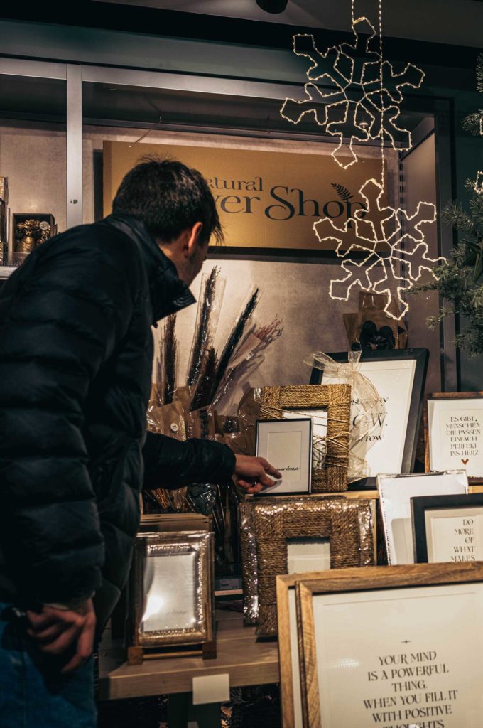 Mann betrachtet Bilderrahen und eingerahmte Sprüche in beleuchtetem Geschäft mit Winterdekoration – stimmungsvolle Streetfotografie von Daniel Lampe