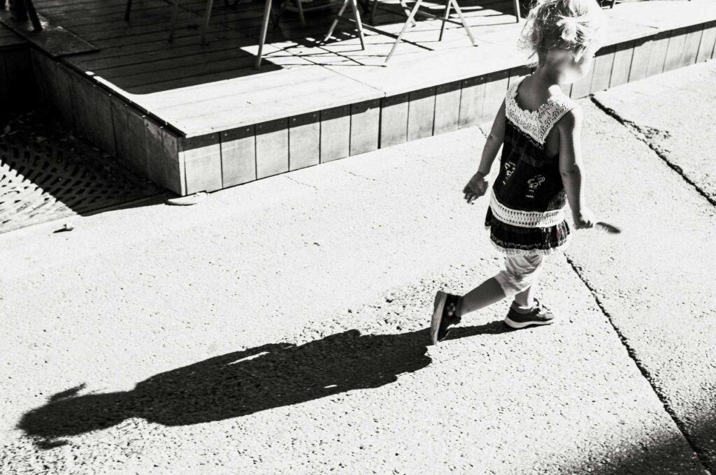Kind läuft in der Sonne über den Asphalt, Schatten fällt auf den Boden – zeitlose Schwarz-Weiß-Streetfotografie von Daniel Lampe