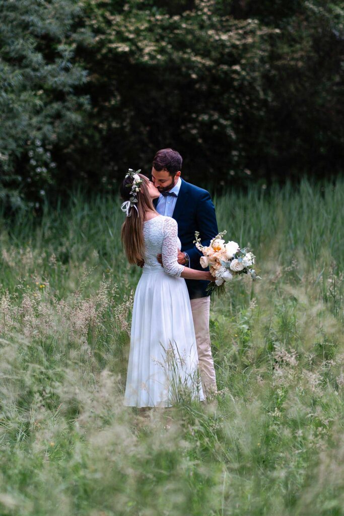 Brautpaar küsst sich in einer Sommerwiese, natürliche Hochzeitsfotografie mit Blumenstrauß und warmem Licht