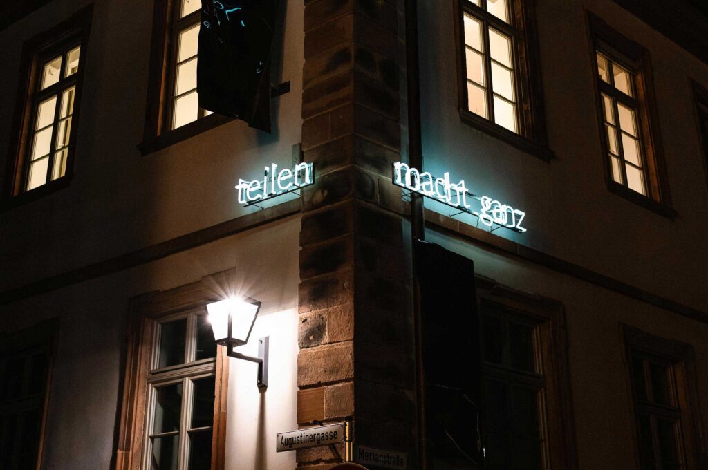 Neon-Schriftzug „teilen macht ganz“ an einer Hausfassade in der Heidelberger Altstadt bei Nacht