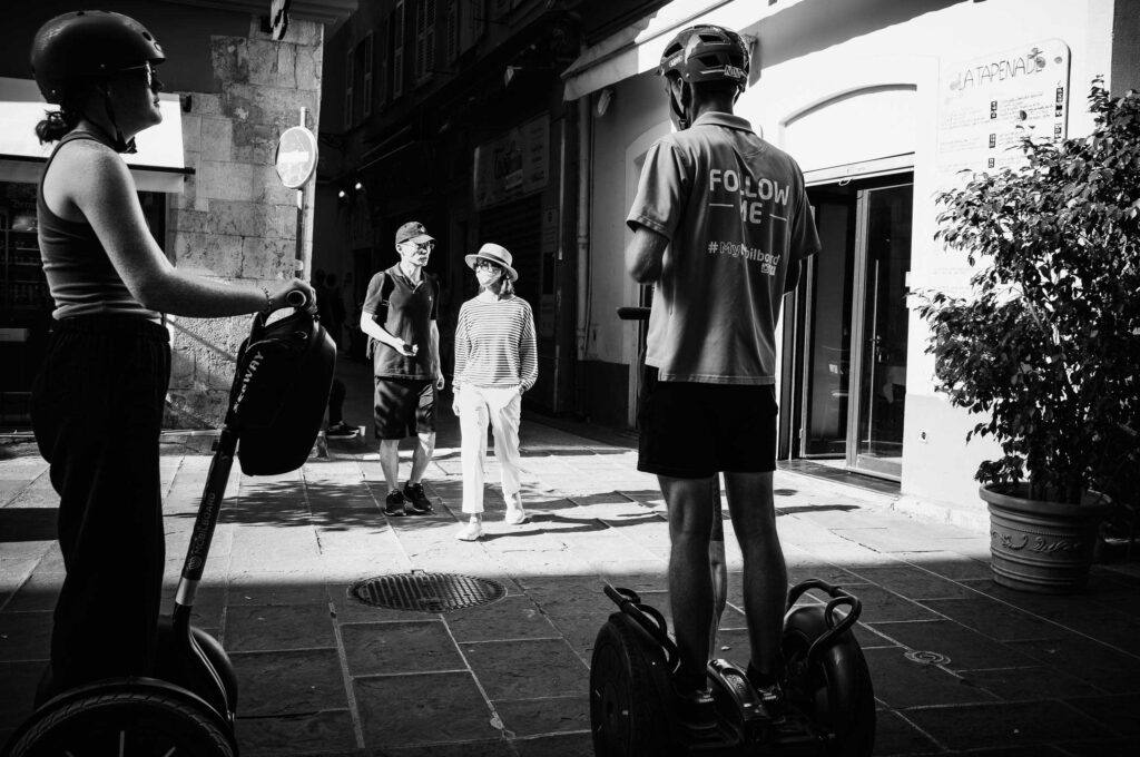 Zwei Segway-Fahrer begegnen einem Paar in den engen Gassen von Nizza – ein Spiel aus Licht, Schatten und urbaner Dynamik.