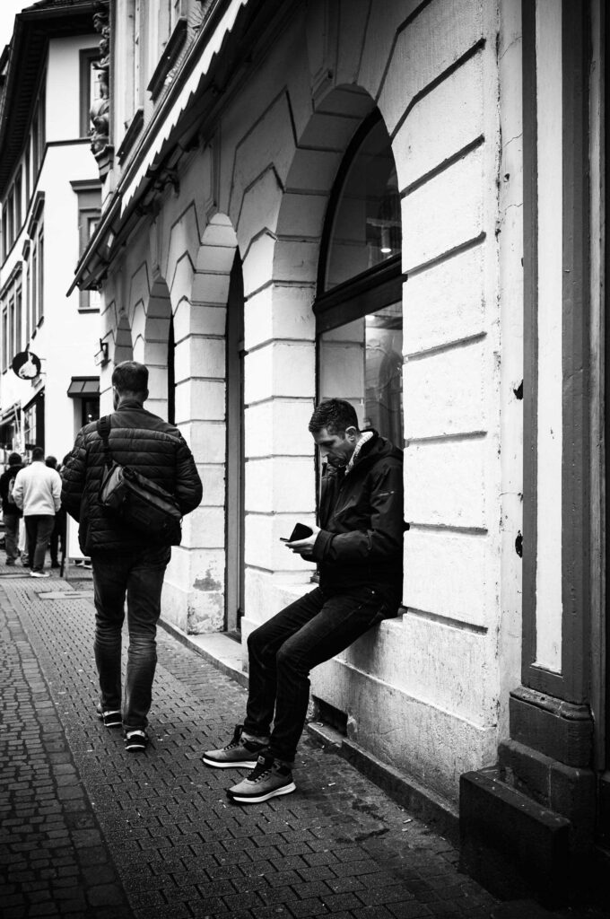 Schwarzweißaufnahme einer Straßenszene: Ein Mann sitzt an einer Hauswand und blickt auf sein Smartphone, während Passanten durch eine historische Altstadtgasse gehen.
