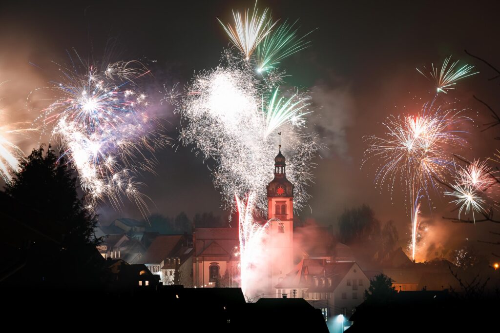 Feuerwerk zu Silvester über einem Dorf mit Kirche bei Nacht, mehrere farbige Explosionen und Lichtspuren vereinen sich zu einer eindrucksvollen Gesamtszene.