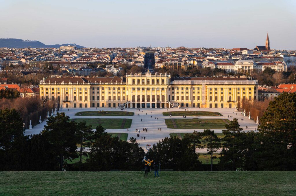 Schloss Schönbrunn in Wien von erhöhtem Standpunkt fotografiert, mit Parkanlage im Vordergrund und Stadtpanorama im Hintergrund bei warmem Abendlicht.