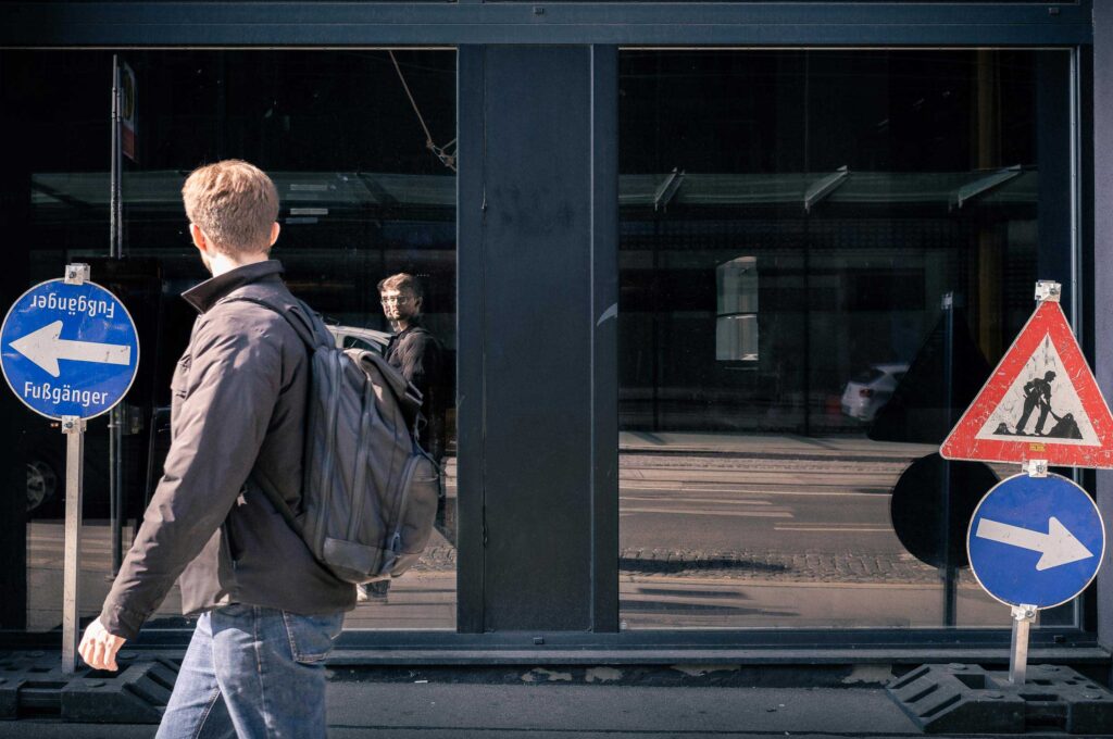 Fußgänger mit Rucksack läuft an Baustellenschildern vorbei, Spiegelung einer Person in Glasfassade in Wien, urbane Szene mit gegensätzlichen Richtungen.